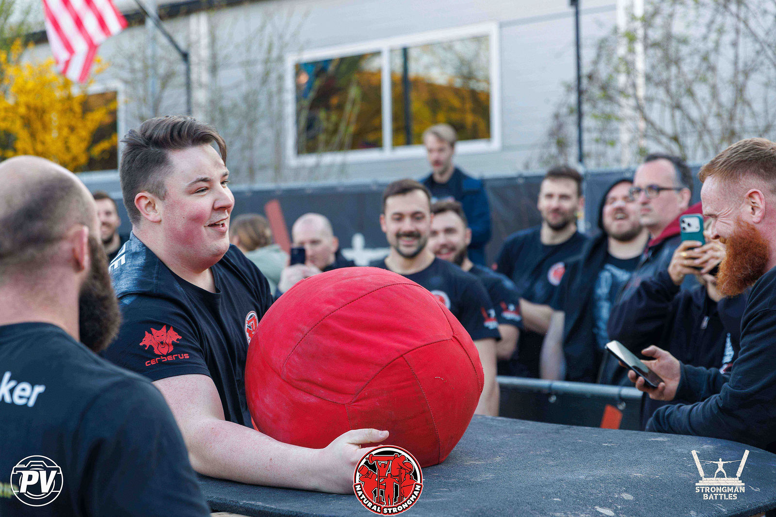 An athlete with a red sandbag at a loading platform, surrounded by supporters.