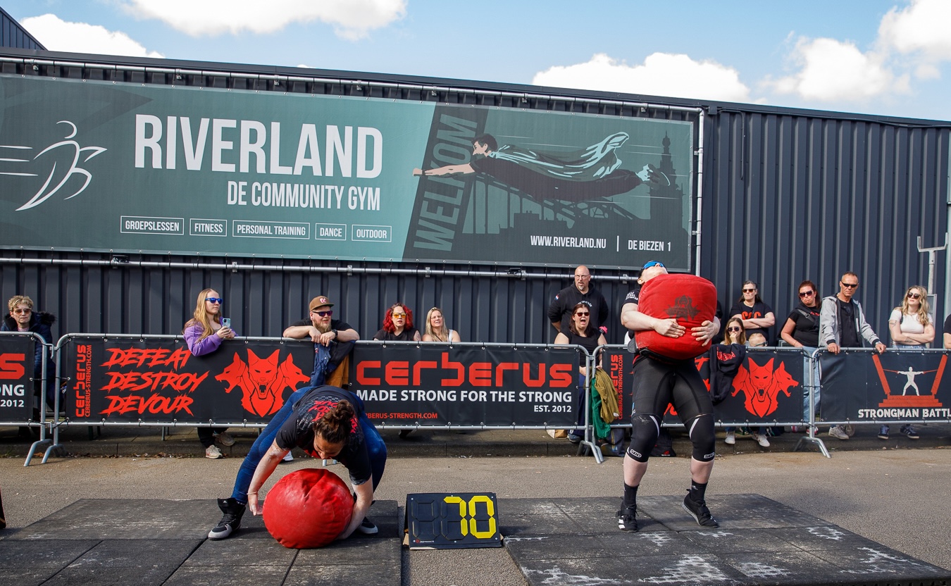 Two athletes compete with sandbags in front of spectators and the Riverland banner at Strongman Battles.